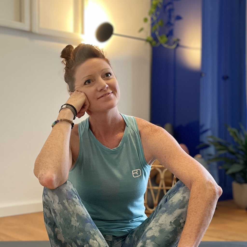 Die Yoganomadin im Portrait, in einem Yogaraum auf ihrer Matte sitzend. Sie lächelt.