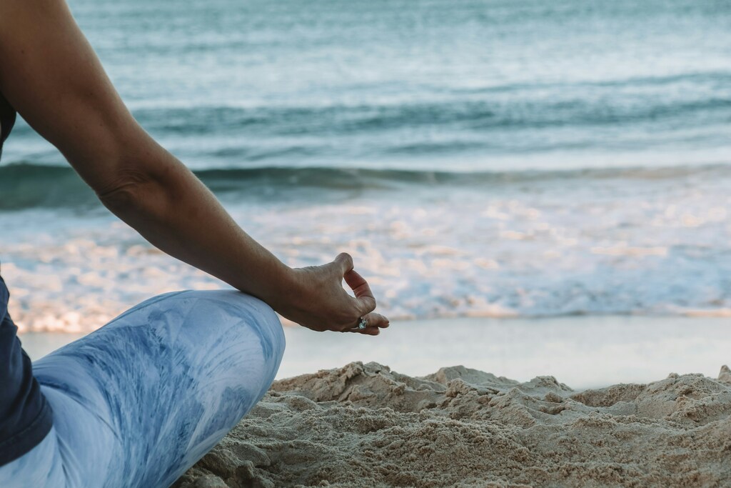 Eine Person sitzt im Schneidersitz im Sitz, die Finger der rechten Hand im Chin-Mudra; im Hintergrund kräuseln sich die Wellen am Strand.